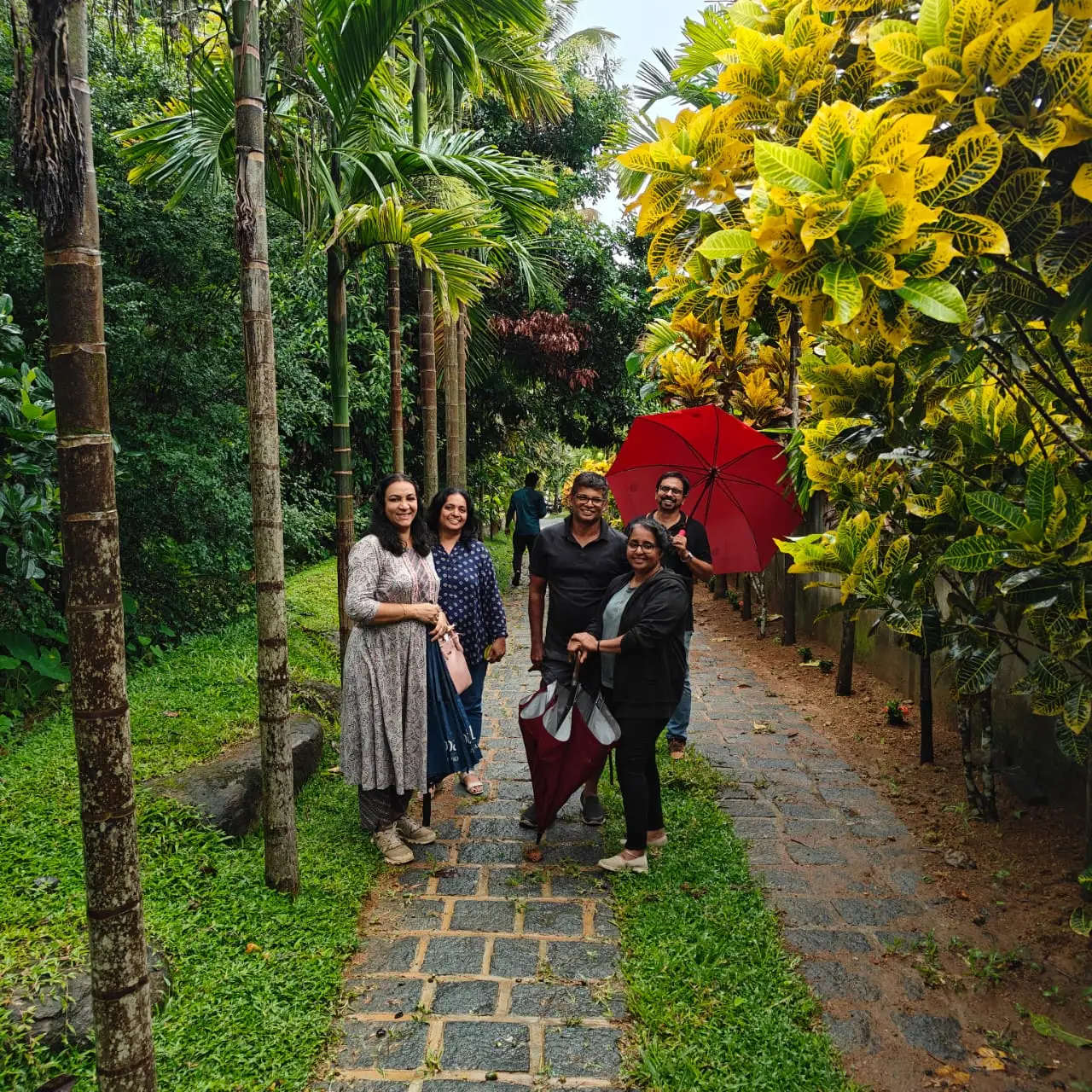 Guests walking the stone garden path at Nyra Thottam beneath areca palms, one carrying a red umbrella