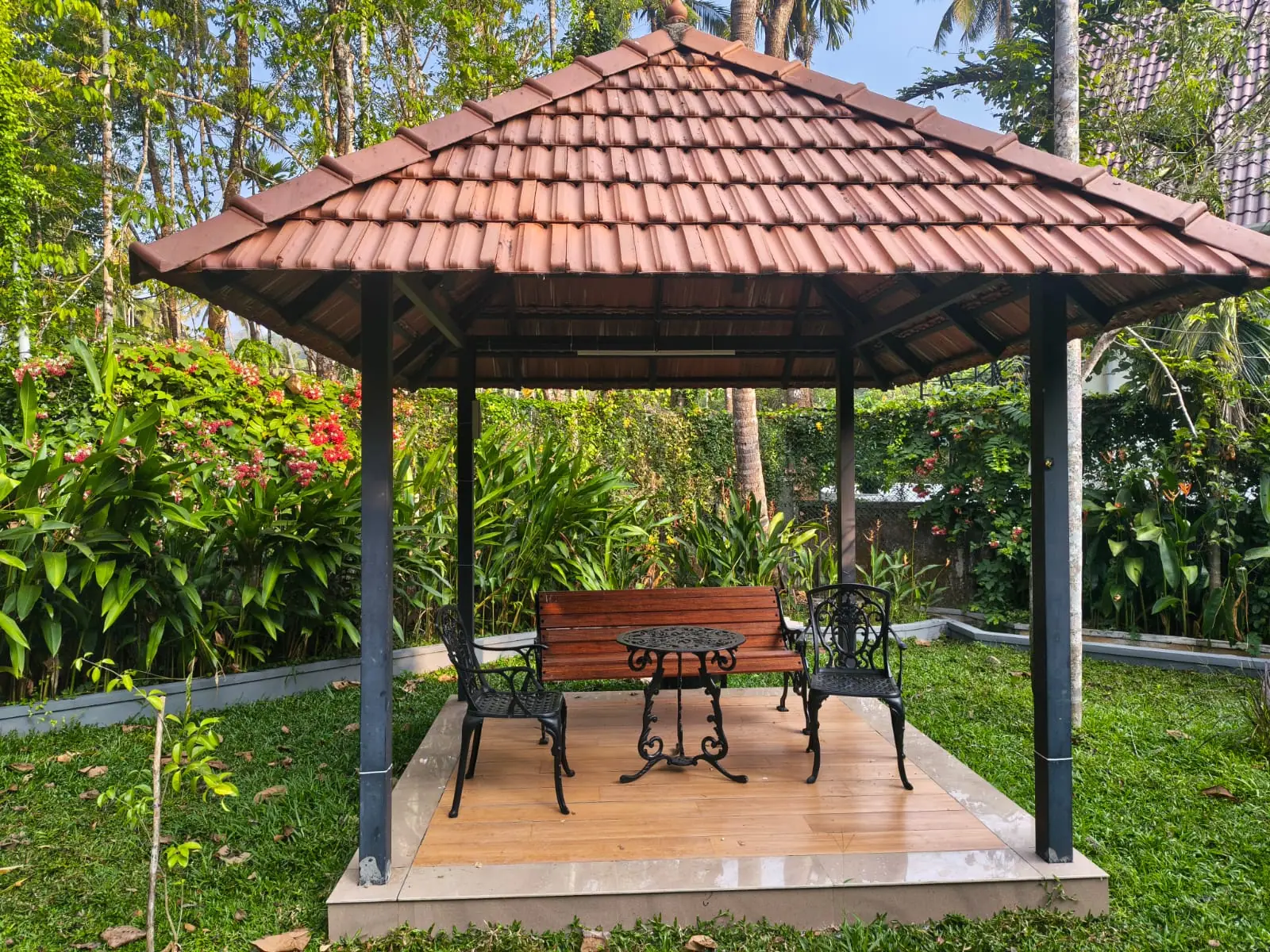 Tile-roofed garden pavilion at Nyra Thottam with a wooden bench and cast-iron table and chairs, surrounded by flowering shrubs