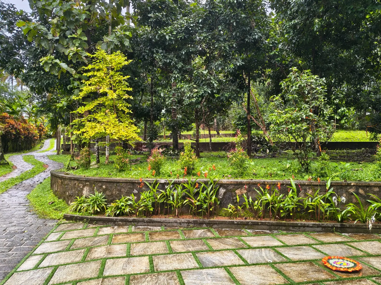 Stone-paved courtyard and curved garden path at Nyra Thottam, edged by raised planted beds glistening after rain